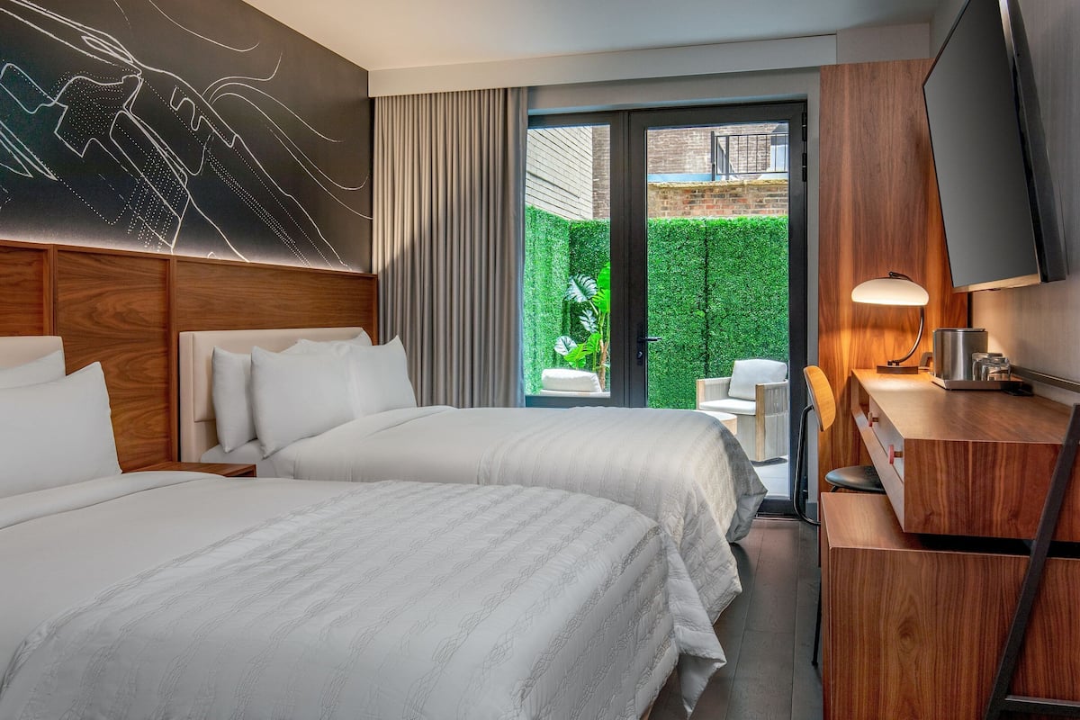 A hotel room with two beds, a TV, a desk, and a view of a green garden.