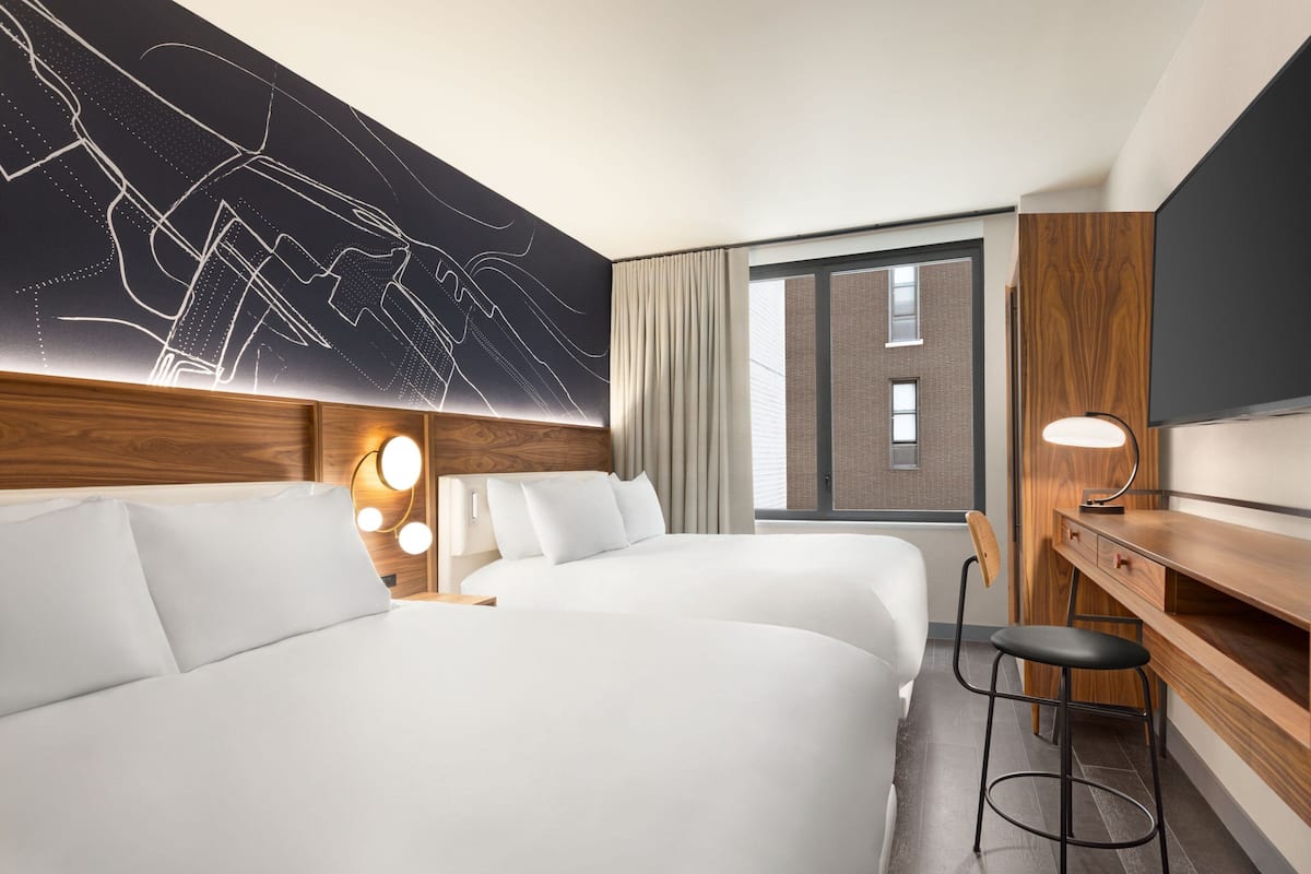 A hotel room with two beds, a desk, a chair, a TV, and a large window with curtains.