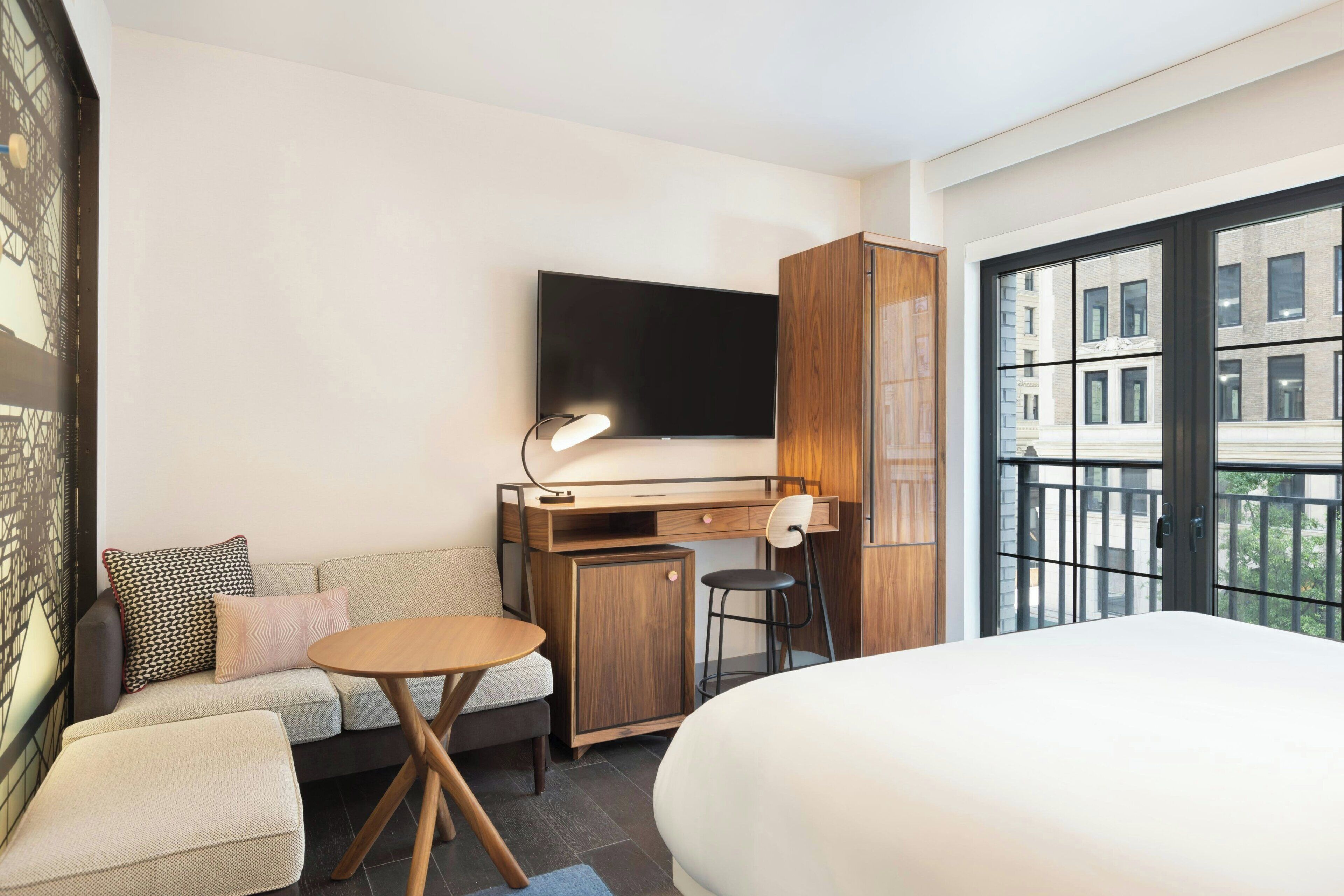 A hotel room with a bed, a desk with a chair, a sofa, a TV, and a balcony with a view of buildings.
