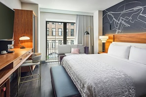 Habitación de hotel con cama, escritorio con silla, banco, ventana grande con vistas a edificios y pared con diseño geométrico.
