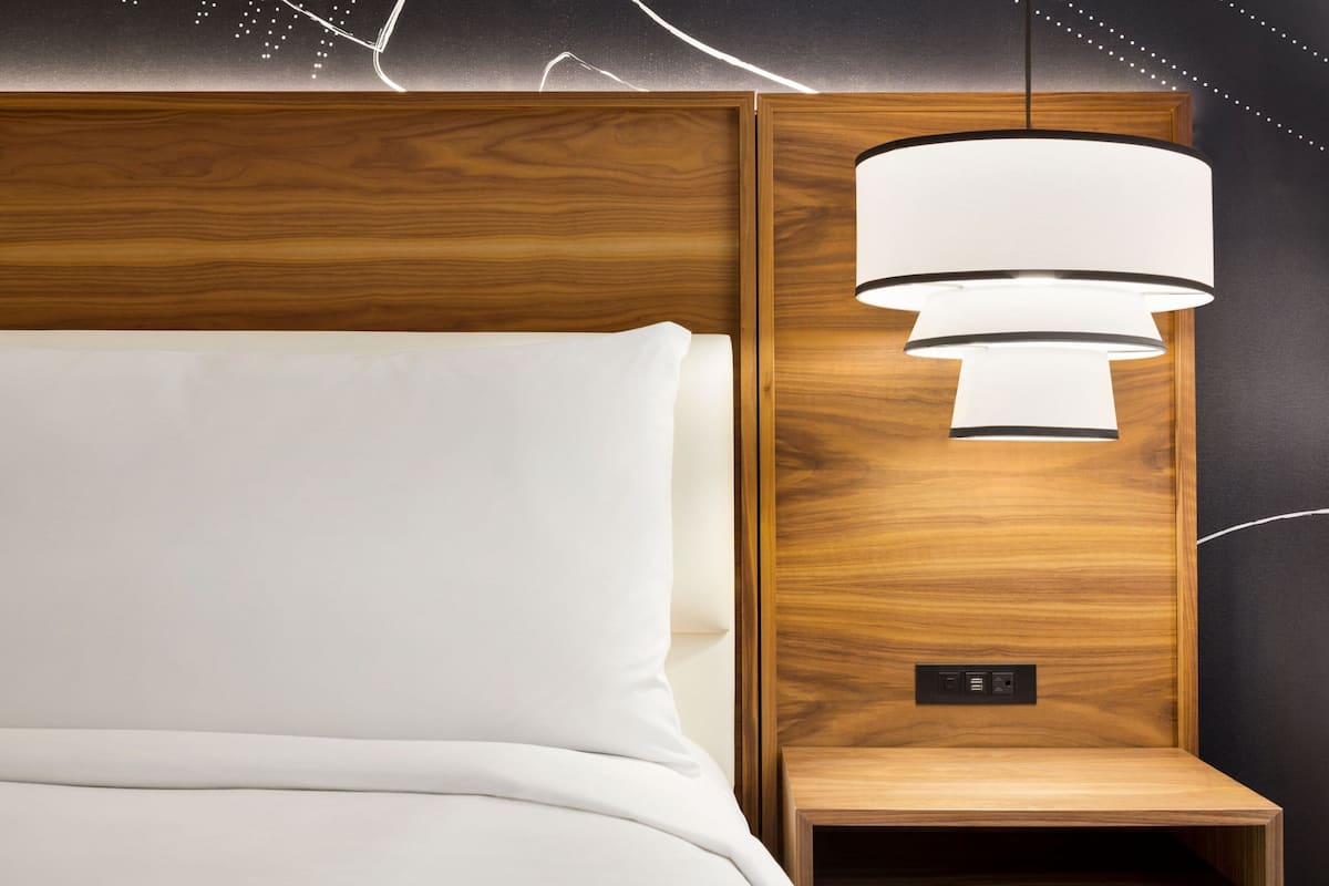 A neatly made bed with white linens, a wooden headboard, and a wall-mounted light fixture.
