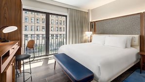 A hotel room with a large bed, a desk with a chair, a bench, a window with curtains, and a view of buildings outside. - Le Meridien New York, Fifth Avenue (New York)