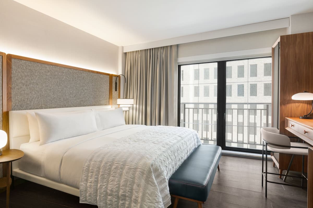 A hotel room with a large bed, a desk with a chair, a bench, and a view of the city through the window.