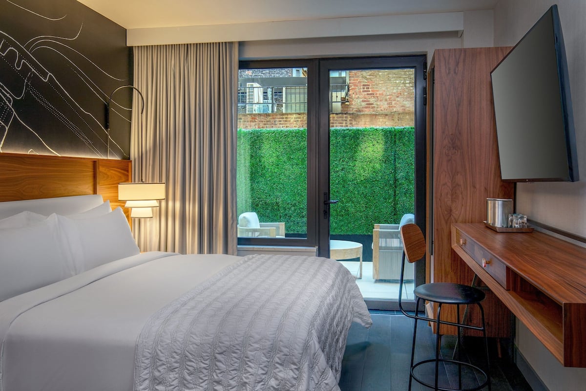 A hotel room with a large bed, a desk with a chair, a TV, and a sliding glass door leading to a balcony with a green hedge and a brick wall.