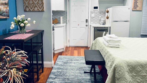 A compact, modern room with a kitchenette, a bed with a neatly folded white blanket, a small table with a vase of flowers, and a bench. - Luxe City Hall Suite by Rittenhouse Square (Philadelphia)