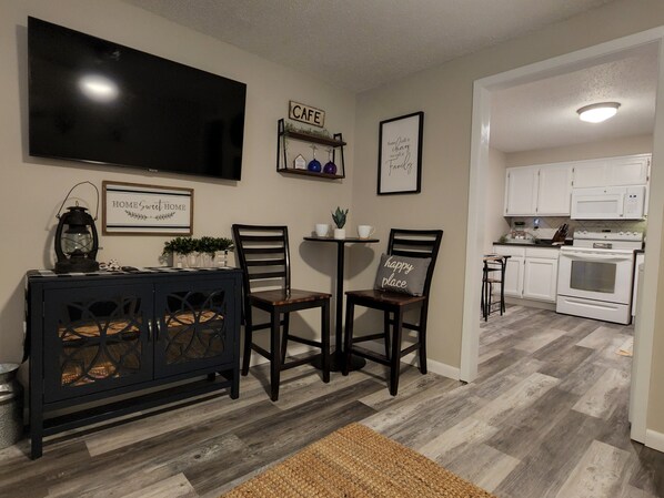 A modern kitchen with white cabinets, a microwave, and an oven. There is a dining area with two chairs and a small table, and a TV mounted on the wall.