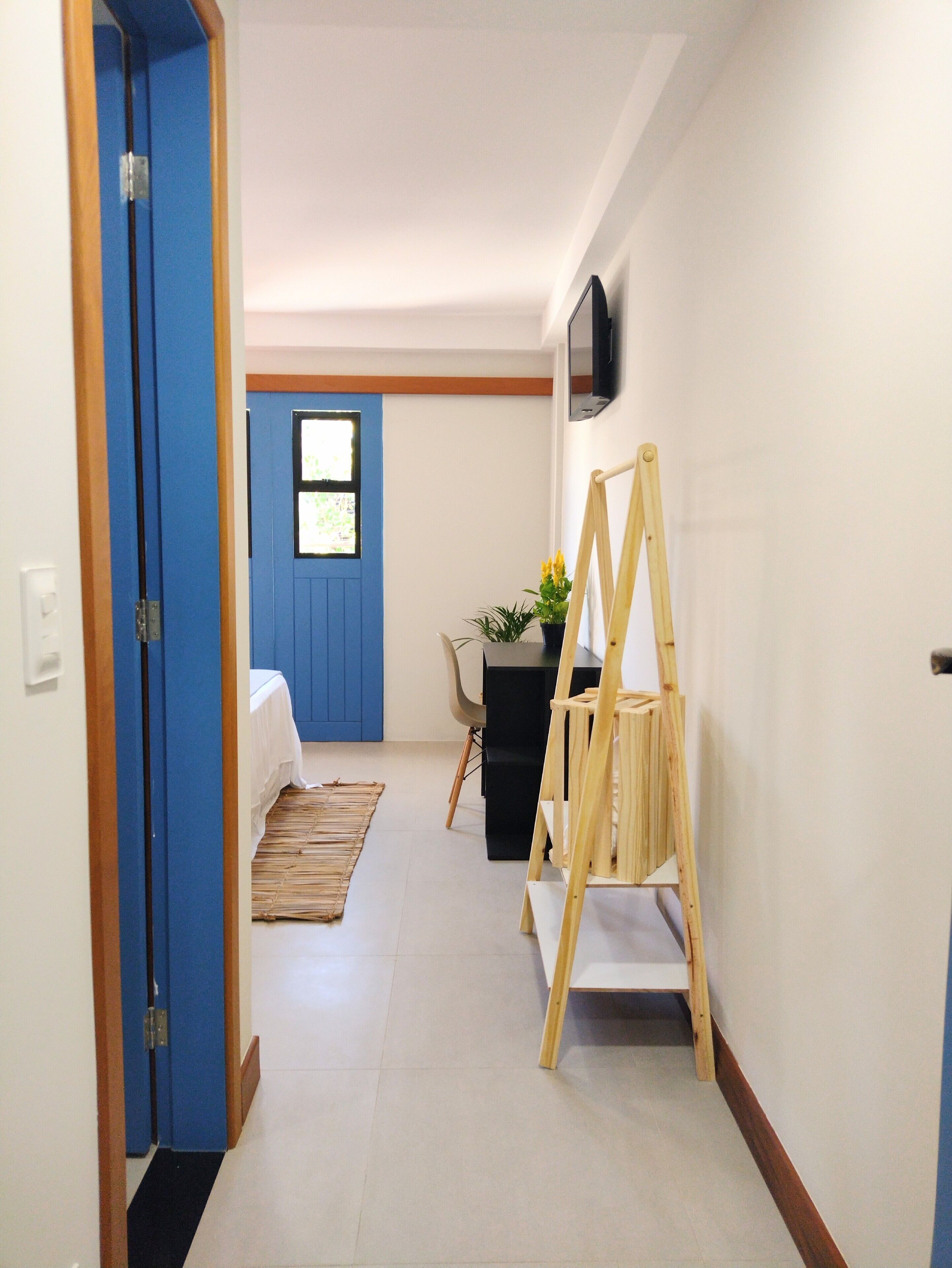 A hallway with a blue door, a bed, a desk with a plant, and a wooden easel.