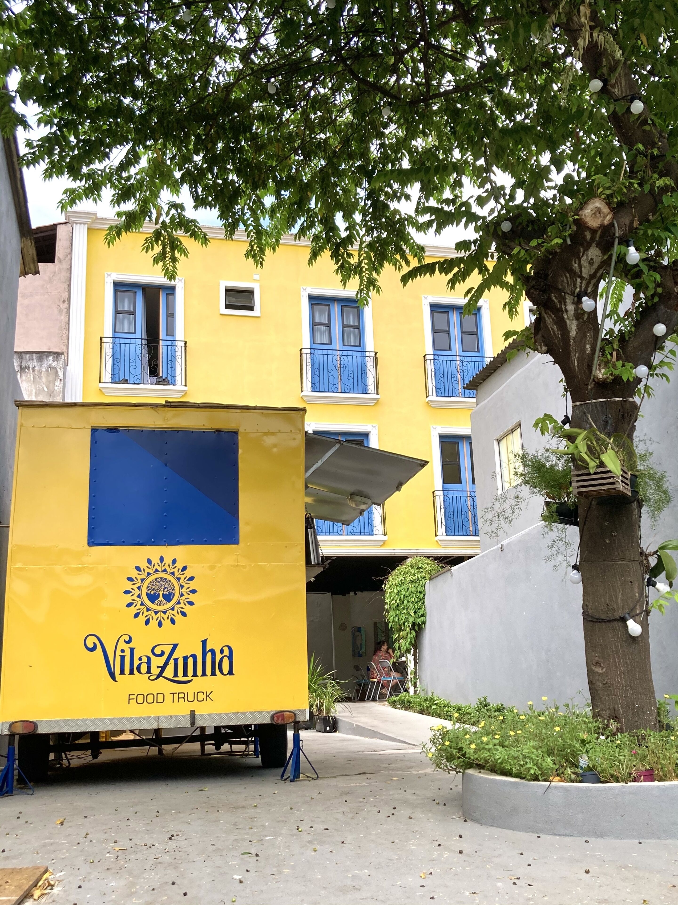A yellow food truck named VilaZinha parked in front of a building with blue balconies.