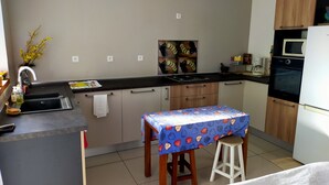 Una cocina moderna con encimera negra, armarios de madera, microondas y horno. Hay un pequeño comedor con una mesa cubierta con un mantel estampado de frutas y dos taburetes.