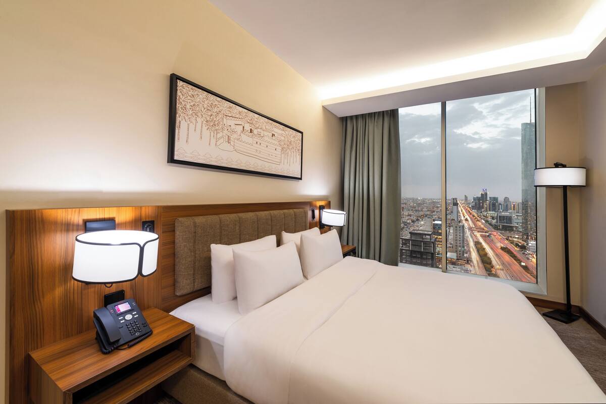 A hotel room with a large bed, a nightstand with a phone, a wall-mounted lamp, and a city view through large windows.