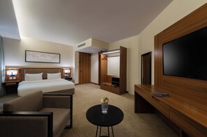 A hotel room with a bed, a desk, a TV, a chair, a small table with a vase, and a wardrobe. - HDB HOTEL- FINANCAL DISTRICT (Riyadh)