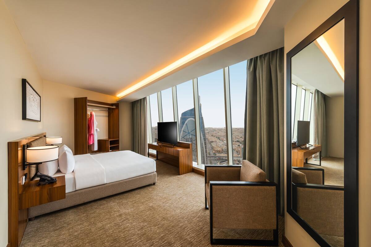 A hotel room with a bed, a TV, a desk, a chair, and a large window with a city view.