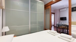 Ein Hotelzimmer mit einem Bett, einem Schreibtisch und einem Sessel. An der Wand befindet sich ein Fernseher und ein Fenster mit Jalousien.