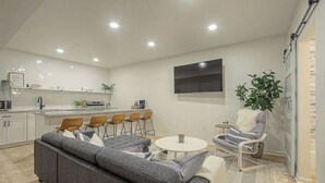 Uma sala de estar moderna com um sofá cinzento, uma mesa de centro redonda e uma poltrona listrada. Uma área de cozinha com armários brancos e uma mesa de jantar com cadeiras é visível ao fundo.