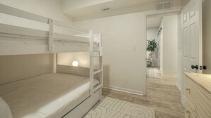 Quarto com beliche, luminária de cabeceira, cômoda de madeira e porta que dá para outro quarto.