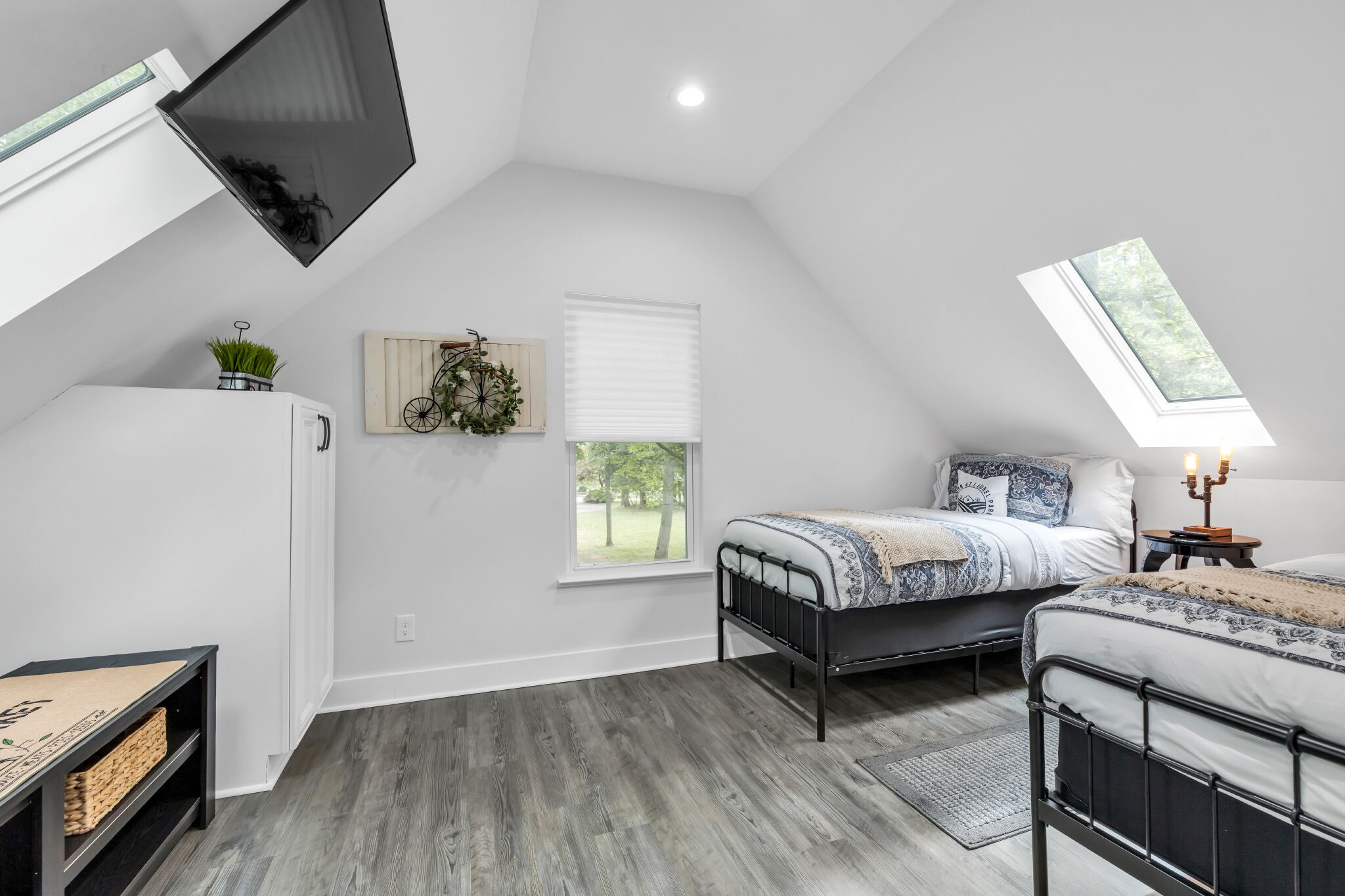 Une chambre avec un plafond en pente, un lit, une table de chevet avec une lampe, une fenêtre et un téléviseur fixé au mur.