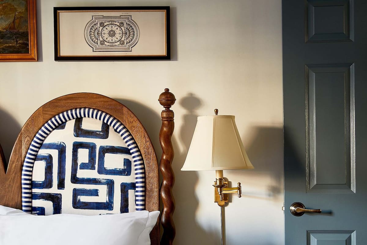 A neatly made bed with a patterned headboard, a bedside lamp, a framed picture above the bed, and a closed door.