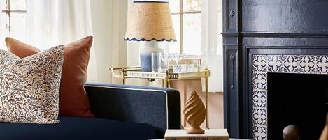 A cozy living room with a dark blue sofa, a round coffee table with books, a lamp, and a fireplace with patterned tiles.