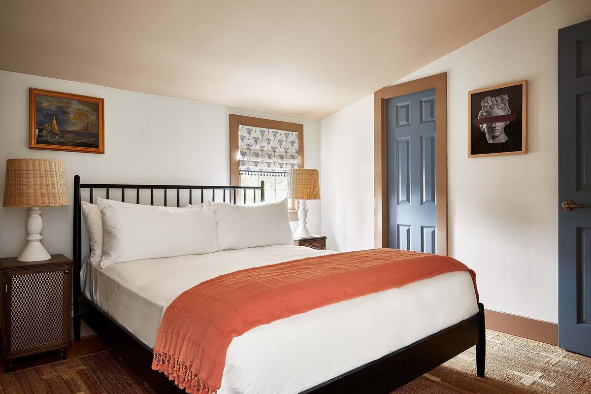 A neatly made bed with white linens and a red throw blanket, a wooden nightstand with a lamp, a window with patterned curtains, and a blue door.