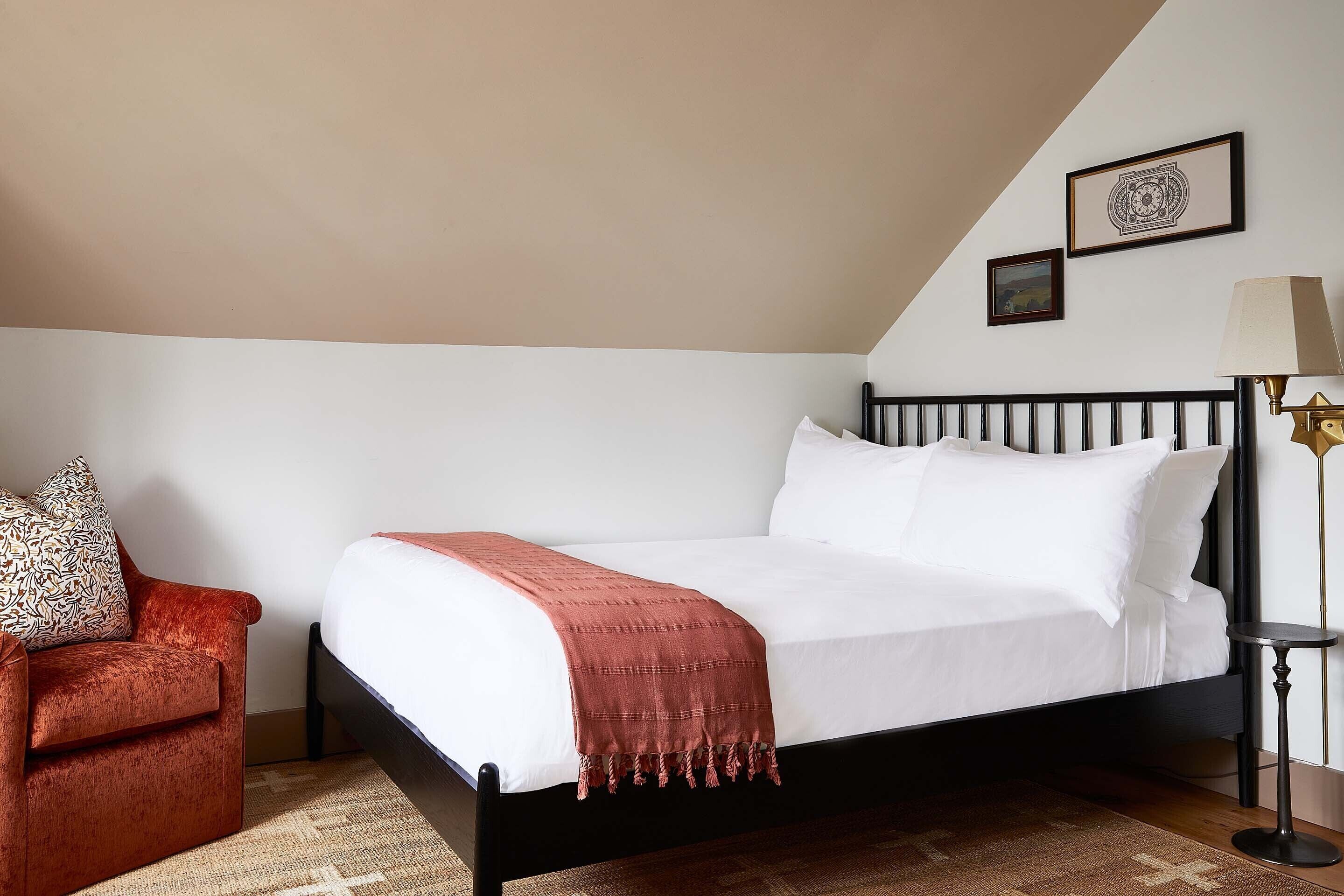 A neatly made bed with a white comforter and a red throw blanket, a red velvet chair, a small round table with a lamp, and two framed pictures on the wall.