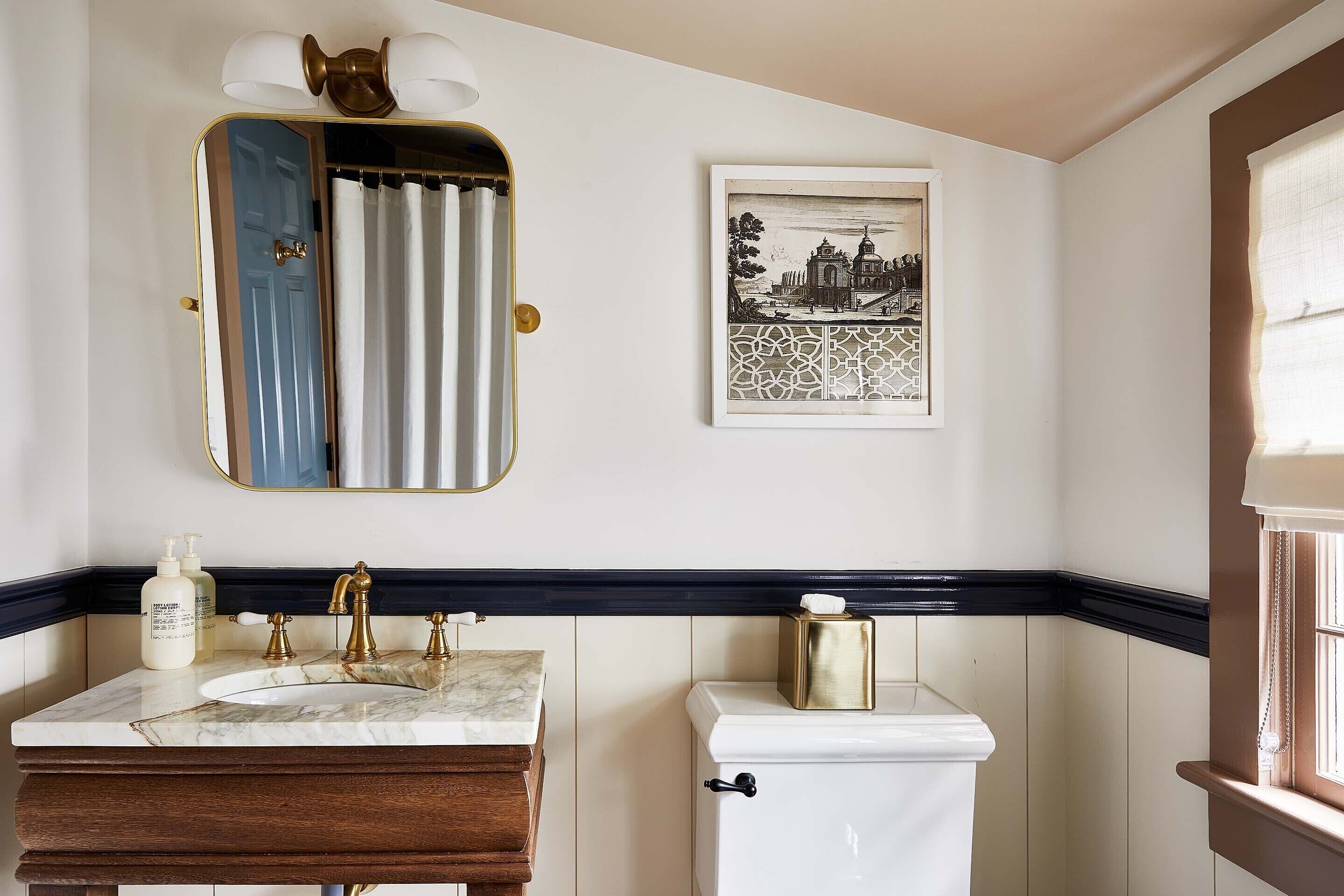 A bathroom with a marble sink, gold fixtures, a toilet with a gold lid, a framed picture on the wall, and a window with a curtain.