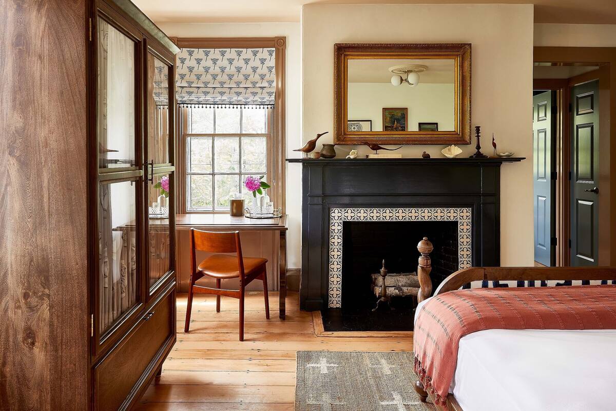 A traditional room with a fireplace, a bed, a desk with a chair, and a window with patterned curtains.