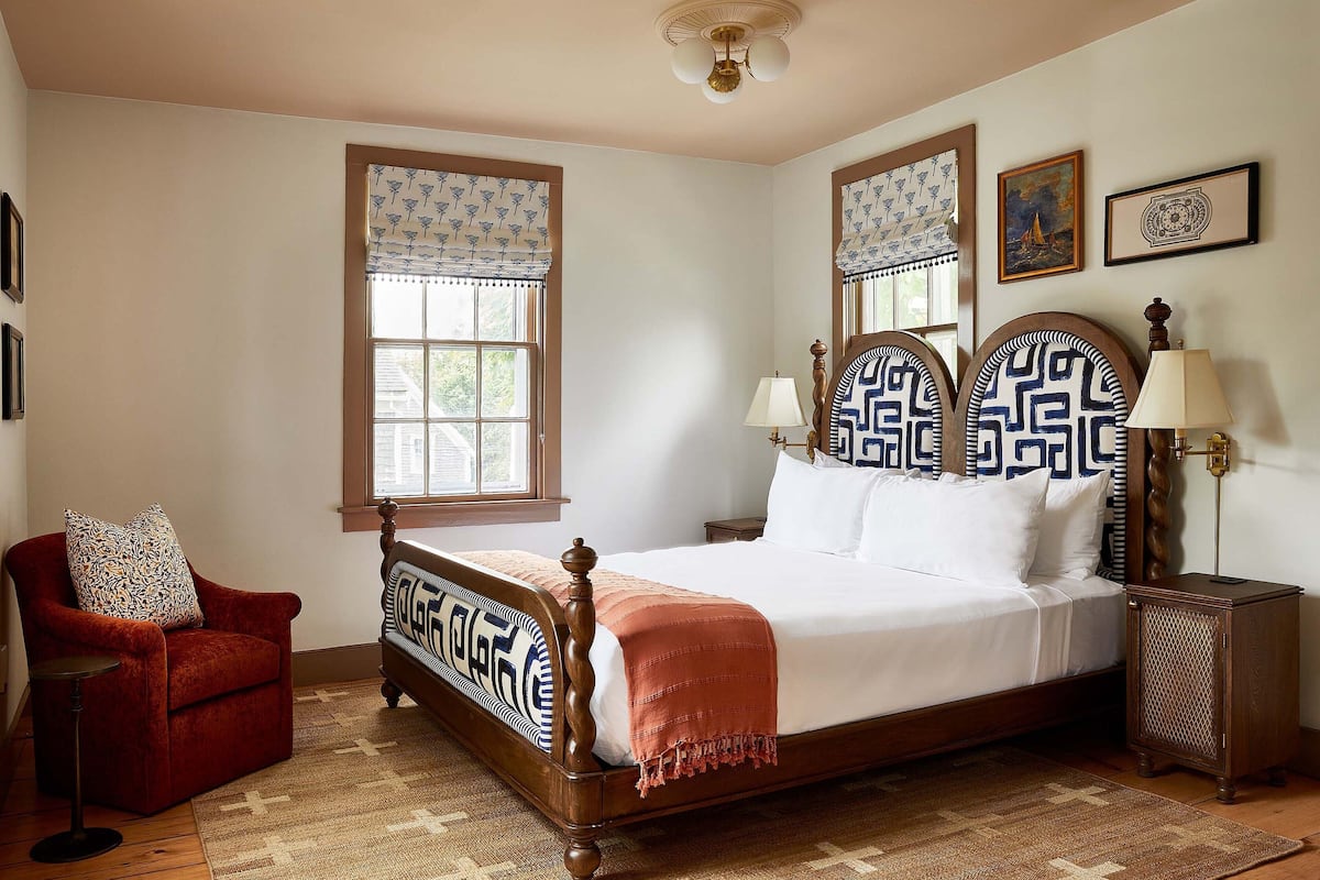 A bedroom with a wooden bed, a red armchair, two bedside tables with lamps, and patterned window valances.
