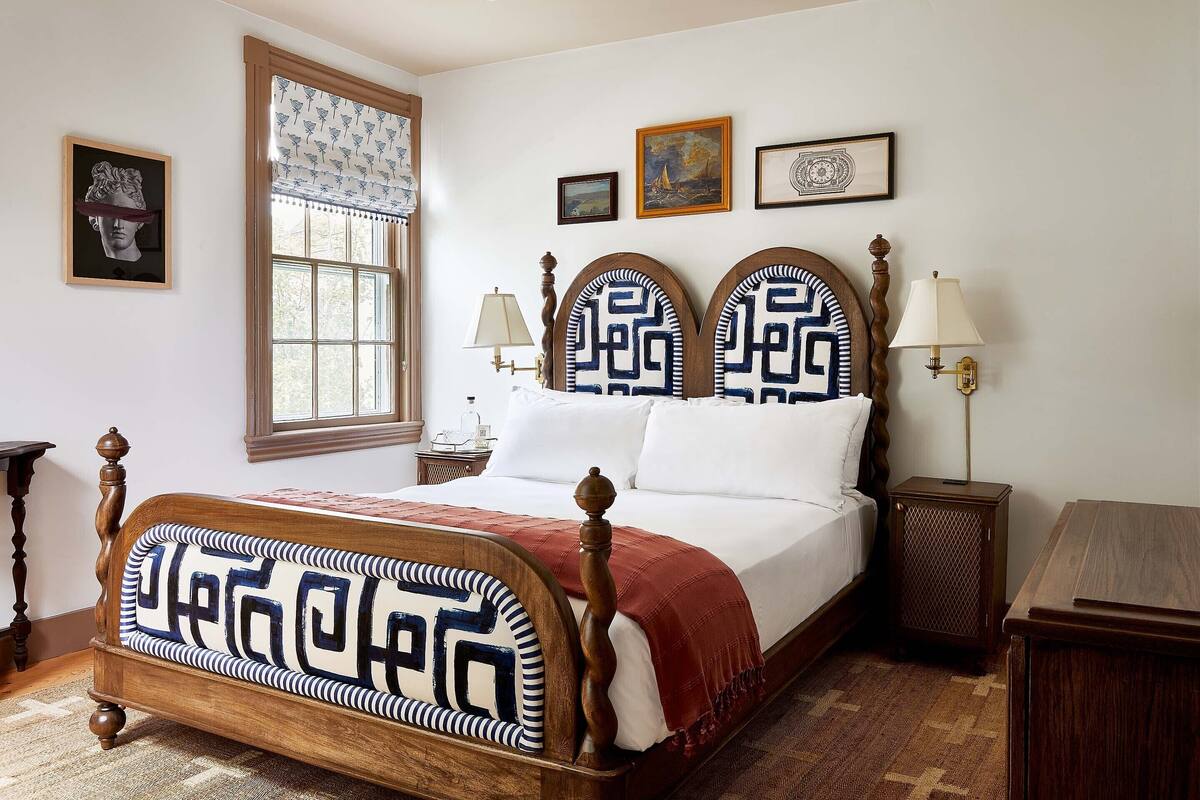 A neatly made bed with a wooden headboard, a window with patterned curtains, and a wall with framed artwork.