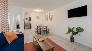 Un salón con sofá azul, una mesa de centro de madera, una zona de comedor con mesa y sillas, una cocina al fondo y un televisor grande montado en la pared.