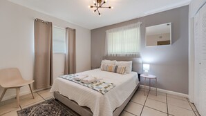 Un dormitorio con cama, una silla, mesita de noche, lámpara y ventana con cortinas.