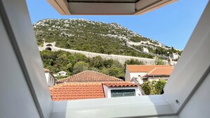 Vista desde una ventana con vista a una montaña, un muro de piedra y edificios con tejados rojos.