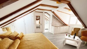 Un dormitorio con una cama grande, una mesita con una botella y una silla. Se ve una cocina y un baño a través de una puerta abierta.