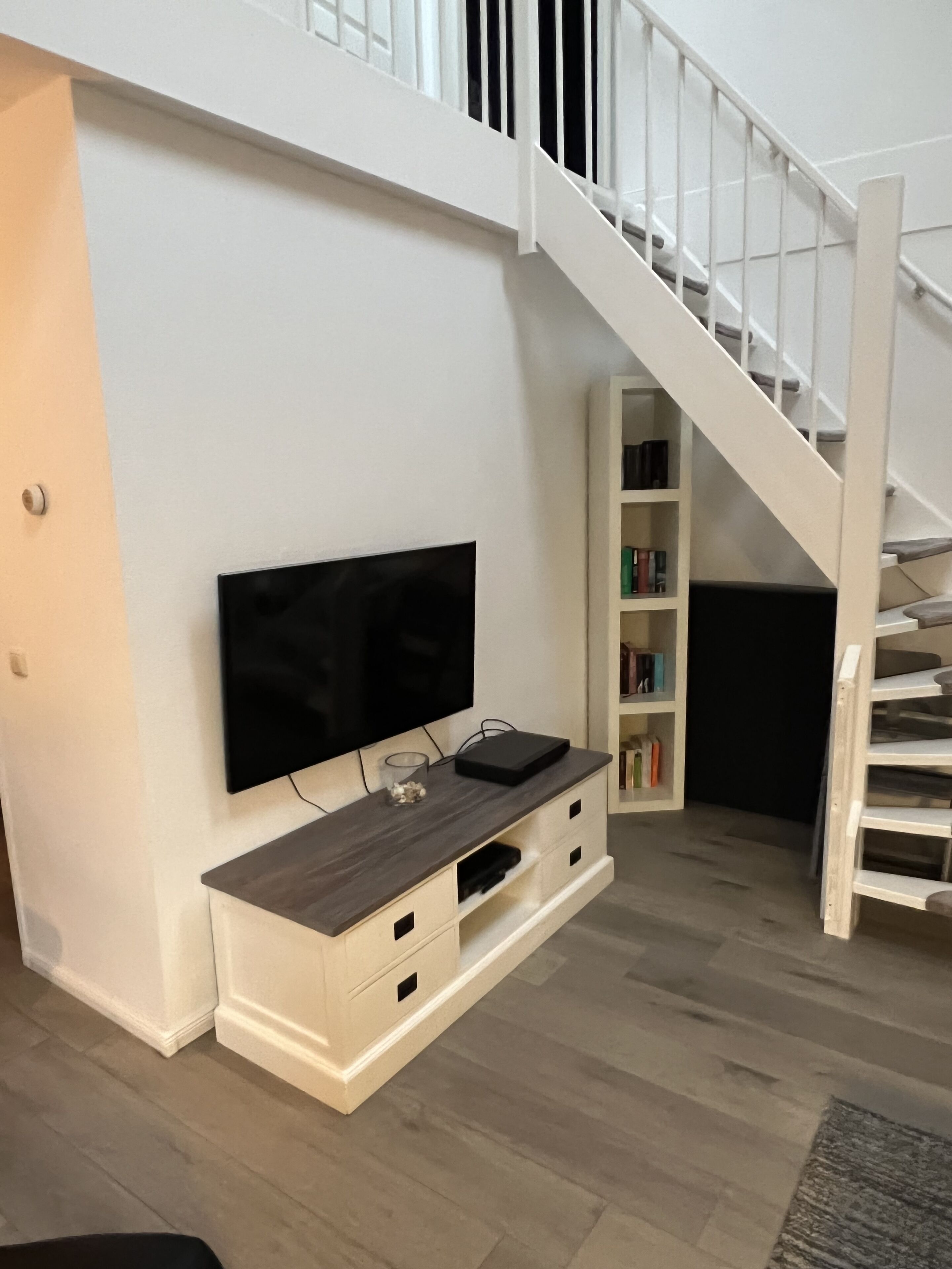 Una sala moderna con un televisor de pantalla plana montado en una pared blanca, un mueble bajo para equipo multimedia de madera y una escalera que conduce a un piso superior.