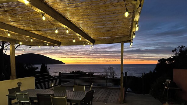Eine Außenpatio mit einer Holzpergola, Lichterketten und Blick auf den Sonnenuntergang über dem Meer.