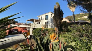 Ein modernes, zweistöckiges Haus mit überdachter Terrasse, umgeben von üppigem Grün und hohen Bäumen.