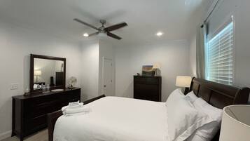 Une chambre à coucher comprenant un lit, une commode, un miroir, un ventilateur de plafond et une fenêtre avec des stores.
