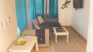 A living room with a sofa, two small tables, a TV mounted on the wall, and a ceiling fan.