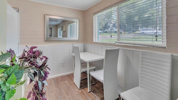 Une pièce compacte et bien éclairée, avec une table et des chaises blanches, une grande fenêtre avec des stores et un miroir au mur.