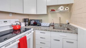 Une cuisine compacte avec des meubles blancs, un plan de travail en granit, une bouilloire rouge, une machine à café, un four à micro-ondes, un évier et une étagère avec du papier toilette et une bouteille.
