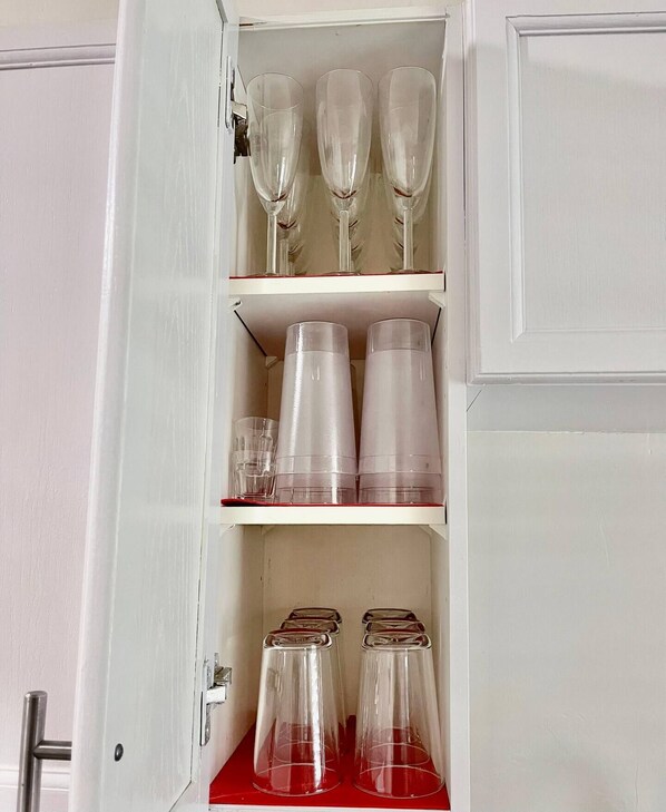 Un armario con tres estantes que contiene copas de vino, vasos de plástico y frascos de vidrio.