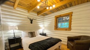 Un dormitorio de cabaña de madera con cama, lámparas de noche, un ciervo montado en la pared, una ventana y un sillón de cuero.