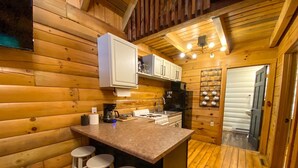 Interior de una cabaña de madera con una zona de cocina que incluye un taburete, una cafetera y un refrigerador.