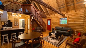 Interior de una cabaña de troncos con cocina, comedor y sala de estar, que incluye una escalera y vigas de madera.
