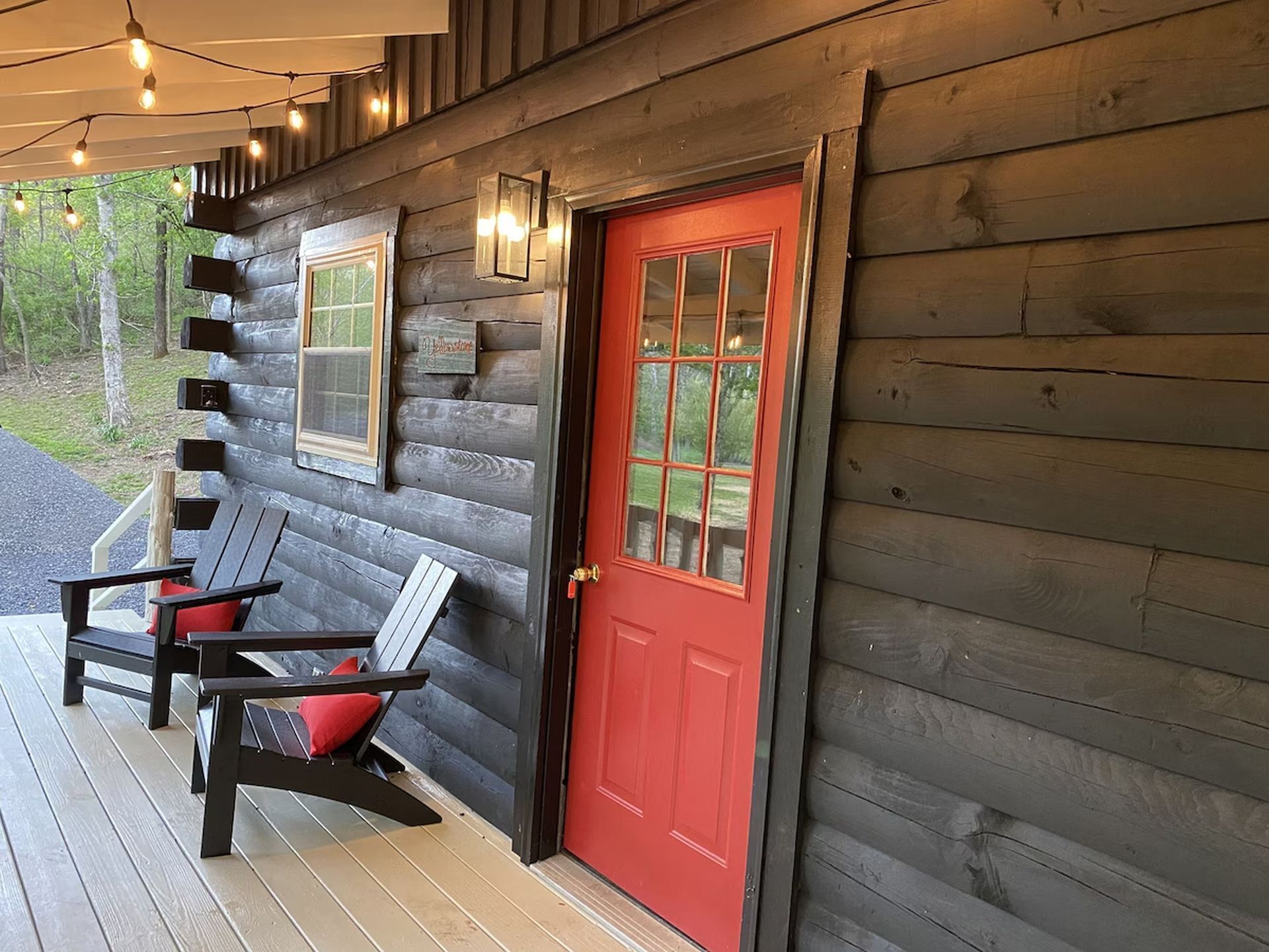 A log cabin with a red door, a porch with two Adirondack chairs, and a sign indicating a nearby attraction.