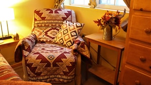 A cozy room with a patterned armchair, a wooden nightstand, a metal vase with autumn leaves, and a window with curtains. - for animal lovers - cute, little Bunkhouse attached to the horse barn (Sonoita)