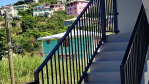 Un escalier avec une rampe bleue mène à un balcon qui donne sur un quartier résidentiel, avec ses maisons aux couleurs vives et sa végétation luxuriante.