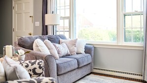 A cozy living room with a grey sofa, patterned cushions, a white lamp, and a large window with a view of greenery outside. - Nona's Cottage-Waynesboro/Shenandoah Valley, VA (Waynesboro)