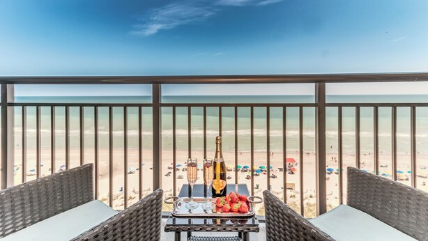 A balcony with wicker furniture, a table with a bottle and glasses, and a view of the beach. - Paradise @ Oceanfront Suite/Dogs OK! (Myrtle Beach)