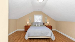 Une chambre avec un plafond en pente, un lit, deux tables de chevet, une fenêtre et un luminaire fixé au mur.