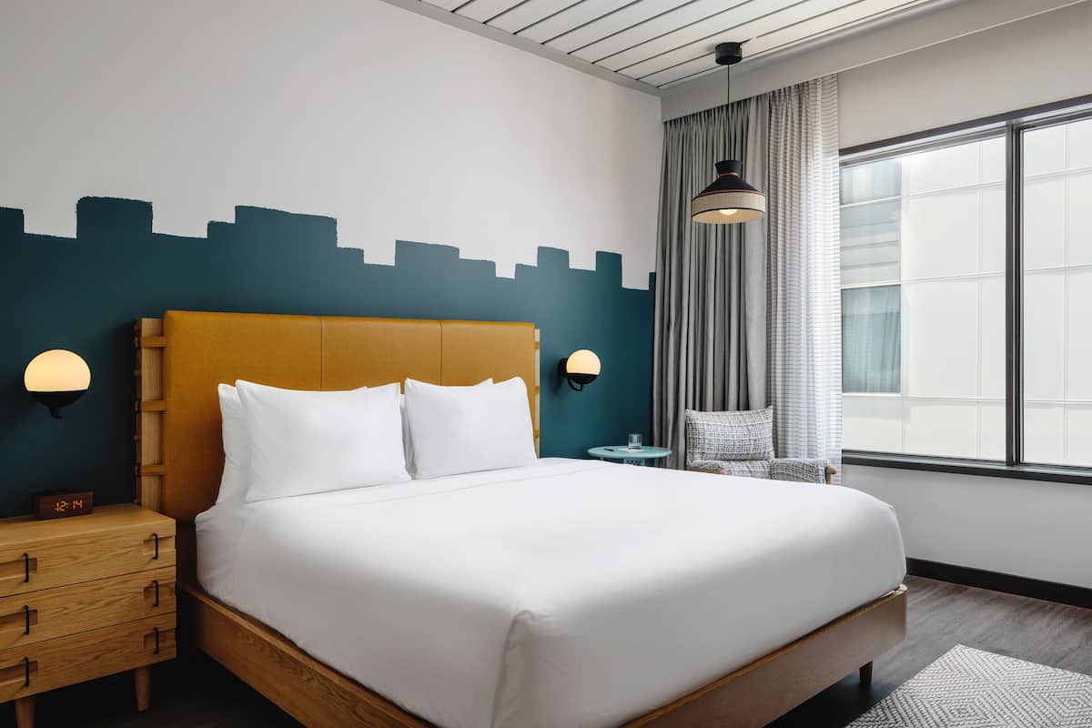 Ein Hotelzimmer mit einem großen Bett, einem hölzernen Kopfteil, zwei Nachttischen mit Lampen, einem Beistelltisch, einem Fenster mit Vorhängen und einer Wandgestaltung mit Stadtansicht.
